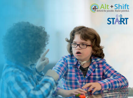 A student wearing glasses and a tracheostomy tube sits at a table, looking toward another child. The students are communicating.